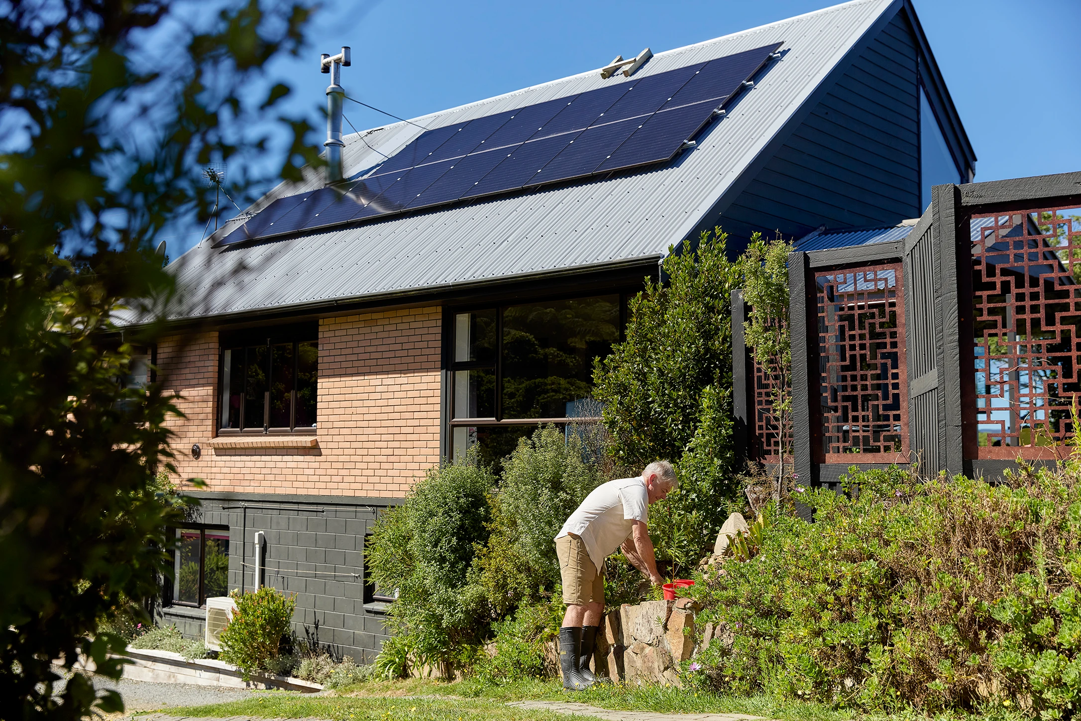 A man is gardening in front of his home, the roof is covered in solar panels.